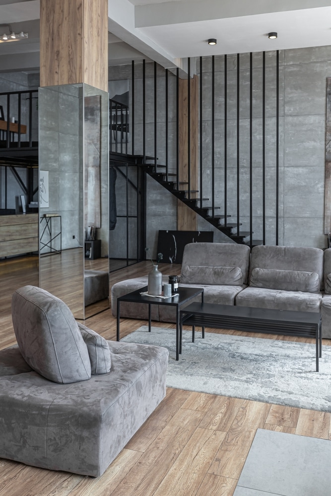 Déco Salon Industriel : Loft Moderne avec Escalier Noir Salon style loft industriel : canapé gris, escalier noir, murs béton et pilier miroir sur parquet clair.