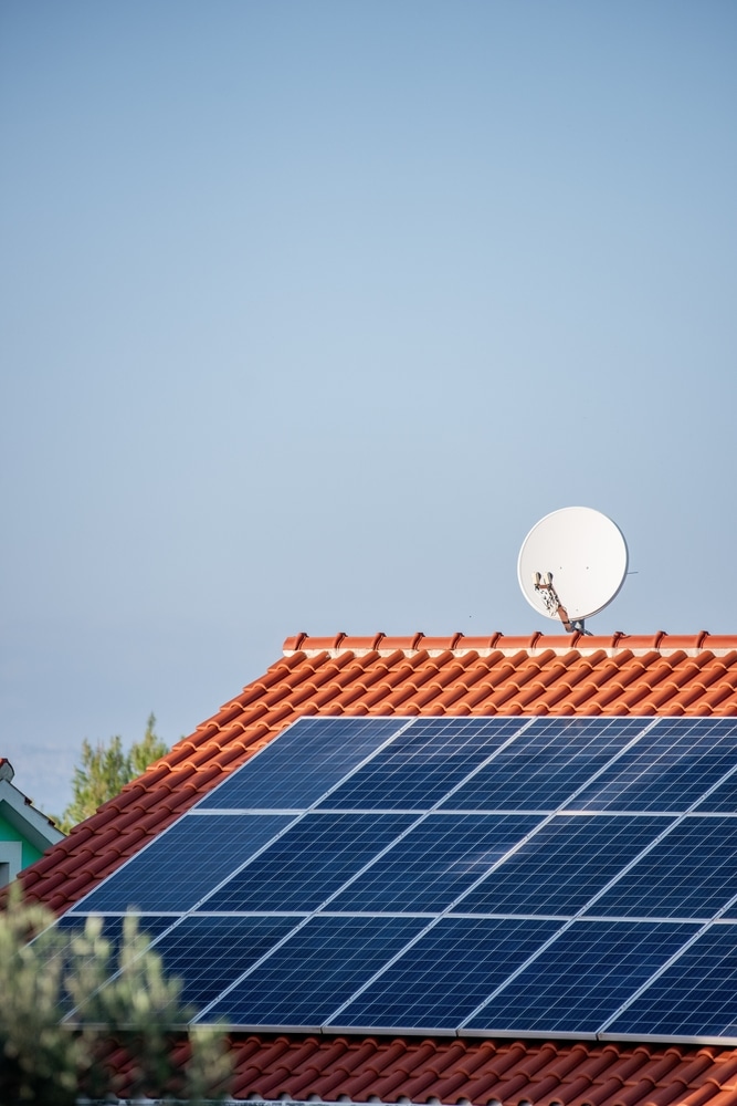 Panneaux solaires et antenne parabolique sur toit de tuiles Panneaux solaires sur toit en tuiles rouges avec antenne parabolique sous un ciel bleu clair dégagé.